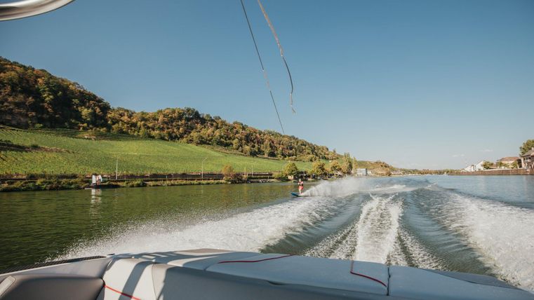 Ein Boot fährt auf einem ruhigen Gewässer, umgeben von sanften Hügeln und grünen Wiesen. Der Himmel ist klar und blau.