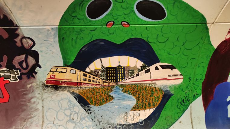 Ein farbenfrohes Wandgemälde zeigt einen großen grünen Kopf mit einem offenen Mund. Aus dem Mund heraus fahren Züge durch eine Stadtlandschaft, die eine kreative Verbindung zwischen Natur und Urbanität darstellt.