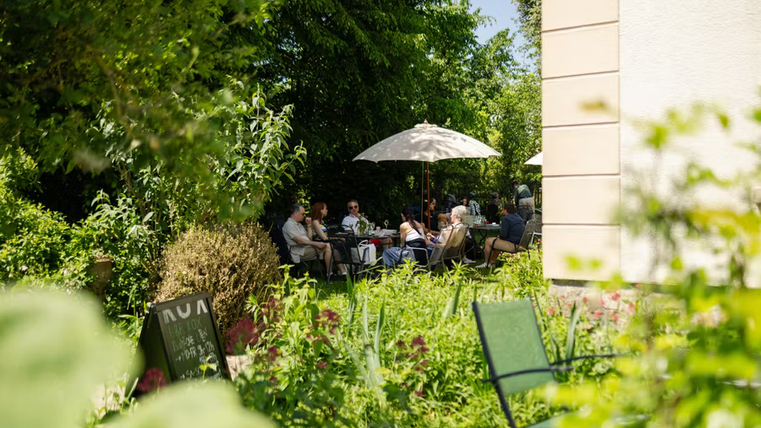 Ein sonniger Garten mit Gästen, die unter Sonnenschirmen sitzen. Umgeben von vielen Pflanzen und einer einladenden Atmosphäre.