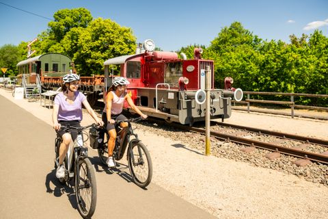 Zwei Radfahrerinnen fahren auf einem Radweg an einer alten Lokomotive vorbei.
