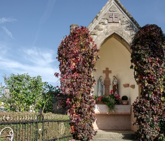 Small chapel with statues, surrounded by autumn ivy. A wrought-iron gate and blue sky in the background., &copy; TI Bitburger Land
