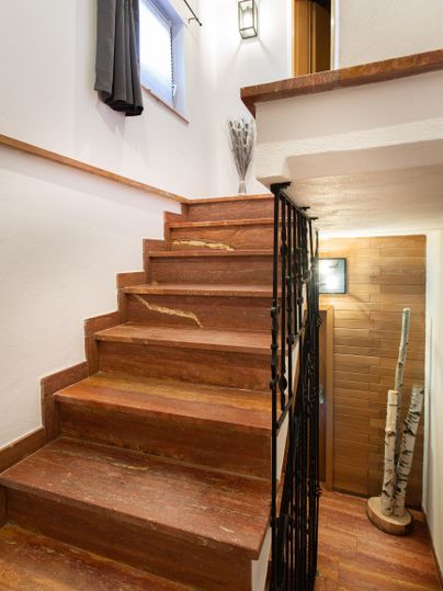 Eine Treppe aus Holz führt nach oben, umgeben von weißen Wänden und einem Fenster mit Vorhängen. Am Ende der Treppe liegt eine Tür, und einige dekorative Elemente sind zu sehen.