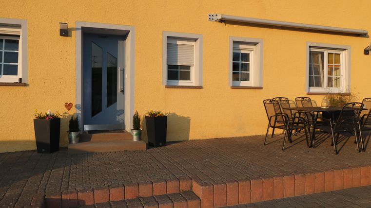 Ein gemütlicher Eingangsbereich mit einer blauen Tür und mehreren Fenstern. Vor der Tür steht ein Tisch mit Stühlen und Töpfen mit Pflanzen.