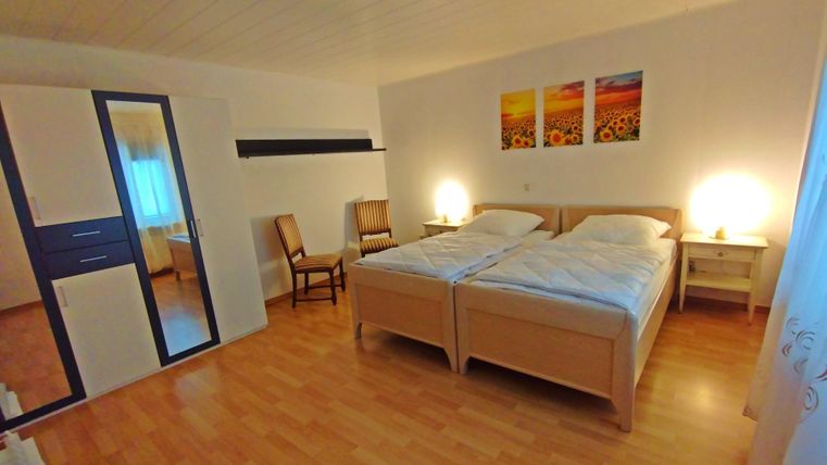 Ein gemütliches Schlafzimmer mit zwei Einzelbetten und warmem Licht. An der Wand hängen Bilder von Sonnenblumen, und es gibt einen Spiegel sowie einen Stuhl.