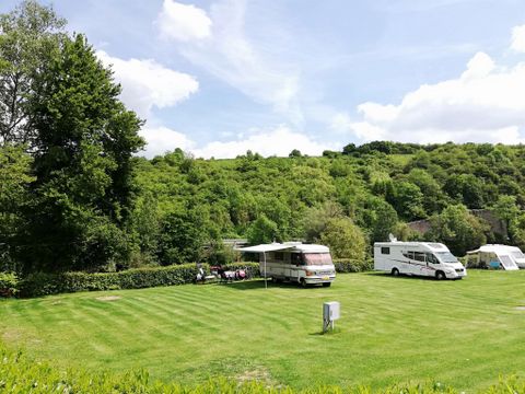 Ein schöner Campingplatz mit mehreren Wohnmobilen auf einer grünen Wiese. Im Hintergrund sind bewaldete Hügel und ein klarer Himmel zu sehen.