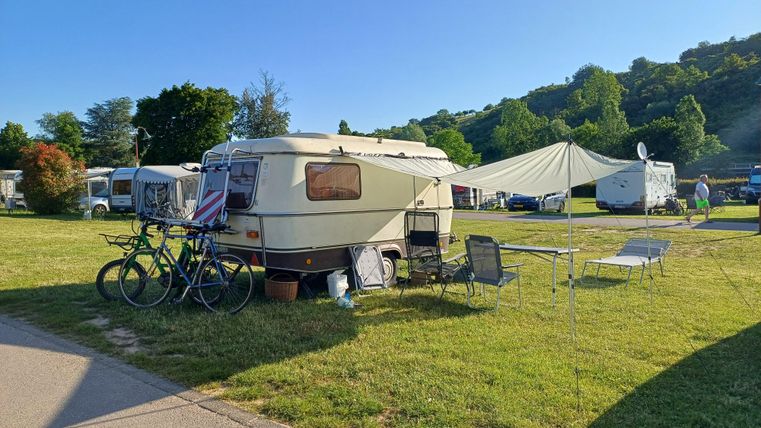 Ein Wohnwagen steht auf einem Campingplatz, umgeben von Bäumen und Wiese. Neben dem Wohnwagen sind Fahrräder und eine kleine Sitzecke zu sehen.