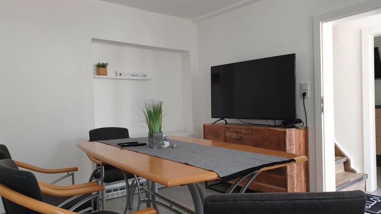 Ein moderner Besprechungsraum mit einem großen Tisch und mehreren Stühlen. An der Wand hängt ein Fernseher, und es gibt Pflanzen zur Dekoration.