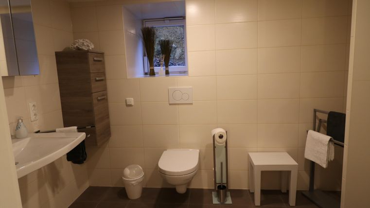 Ein modernes Badezimmer mit hellen Fliesen und einem Fenster. Es enthält ein Waschbecken, eine Toilette und eine kleine Sitzgelegenheit.