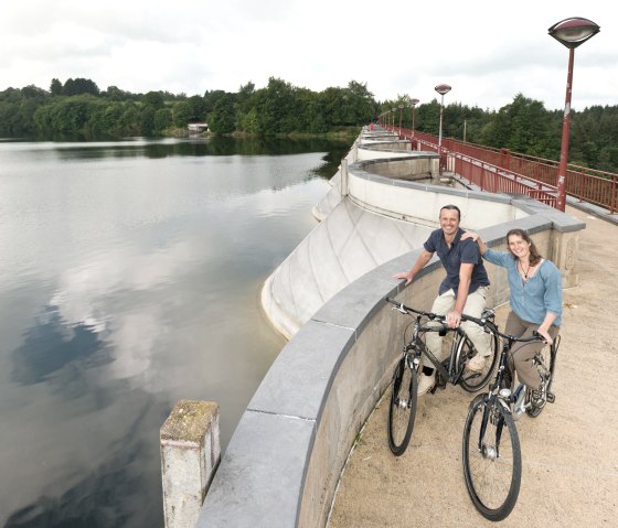 Zwei Personen mit Fahrrädern stehen auf der Staumauer des Stausees Bütgenbach. Im Hintergrund sind Wasser und Bäume zu sehen., © vennbahn.eu