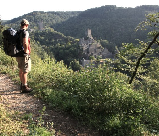 Een wandelaar met rugzak staat op een pad en kijkt uit over de Manderscheid kastelen in het groene heuvellandschap van de Eifel., © Björn Nehrhoff