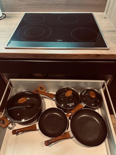 Ein Küchenschrank mit mehreren Pfannen und Töpfen, die Holzgriffe haben. Im Hintergrund befindet sich ein Glaskeramikkochfeld.