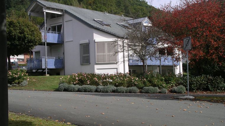 Ein modernes Wohngebäude mit einem angelegten Garten. Im Hintergrund ist ein Hügel und ein blauer Himmel zu sehen.