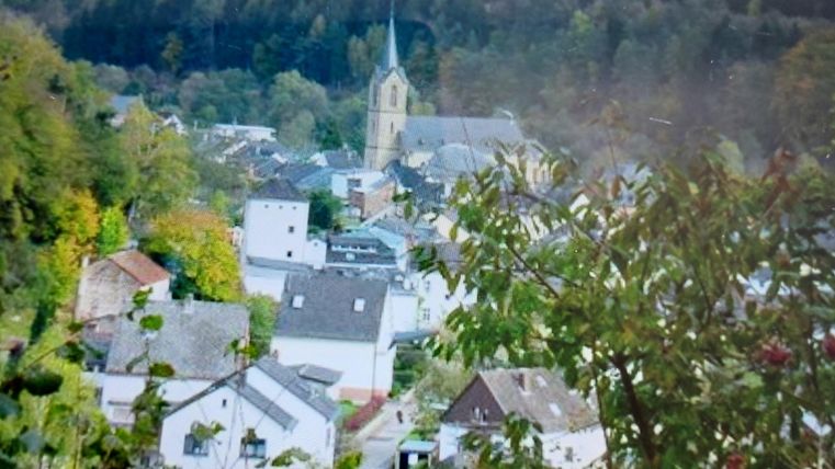 Ein kleines Dorf mit weißen Häusern und einer Kirche im Hintergrund. Umgeben von grünen Bäumen und Hügeln.