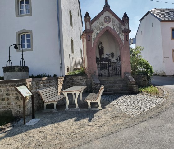 Petite chapelle avec statue et banc à Wolsfeld. On peut également y voir une fontaine et un panneau d'information. Journée ensoleillée., © Tourist-Information Bitburger Land, Steffi Wagner
