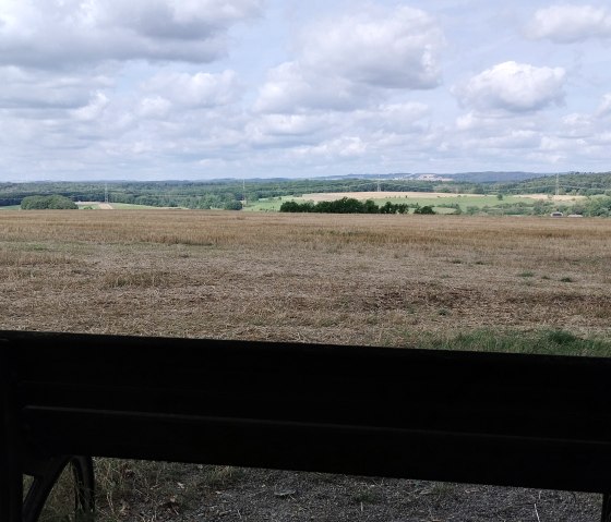 A bench stands in front of a wide field with a view of wooded hills under a cloudy sky., &copy; TI Bitburger Land