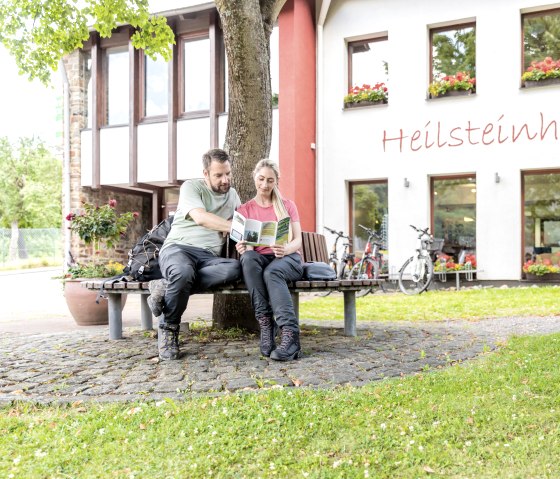 Twee mensen zitten op een bankje voor het Heilsteinhaus in Einruhr en lezen een brochure. Er staan fietsen geparkeerd voor het gebouw, dat is versierd met bloemen., © -@Eifel Tourismus GmbH, AR-shapefruit-AG