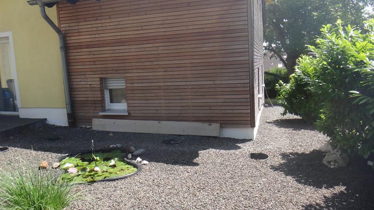 Ein ruhiger Außenbereich mit einer Holzverkleidung und kieseliger Fläche. Auf der linken Seite ist ein kleiner Teich mit Pflanzen sichtbar.