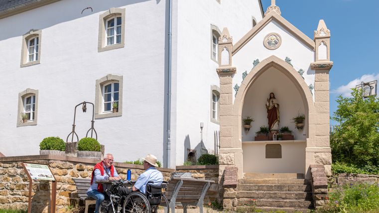 Zwei Männer sitzen auf einer Bank vor einem weißen Gebäude mit einer religiösen Statue in einer Nische.