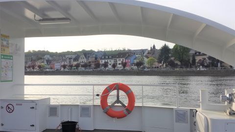 Eine Aussicht von einem Boot auf eine malerische Uferlandschaft. Im Vordergrund ist ein Rettungsring und im Hintergrund sind bunte Häuser und Bäume zu sehen.