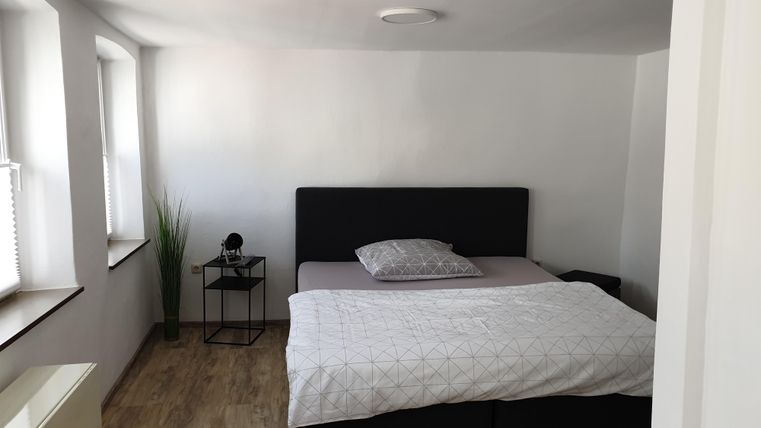 Ein einfaches Schlafzimmer mit einem großen Bett und natürlichem Licht. Eine Pflanze und ein Nachttisch ergänzen die Einrichtung.