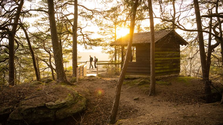 Zwei Personen spazieren bei Sonnenuntergang an einem Aussichtspunkt im Wald vorbei, neben einer Holzhütte.