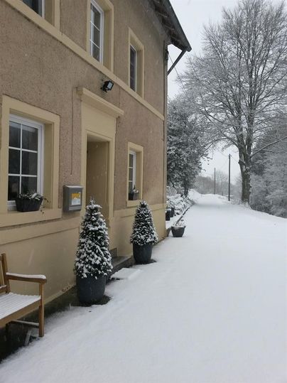 Ein winterliches Szenario mit einem Gebäude, umgeben von Schnee. Die Straße ist ebenfalls schneebedeckt und mit schneebedeckten Pflanzen dekoriert.