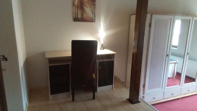Ein schlichtes Zimmer mit einem Schreibtisch und einem Stuhl. Im Hintergrund befindet sich ein Kleiderschrank und ein Bild an der Wand.