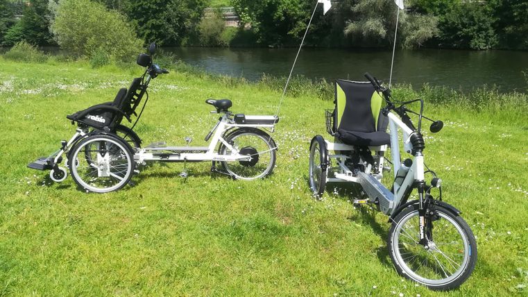 Zwei besondere Fahrräder stehen auf einer grünen Wiese neben einem Fluss. Das eine Fahrrad ist ein Liegedreirad und das andere ein elektrisch betriebenes Zweisitzerfahrrad.