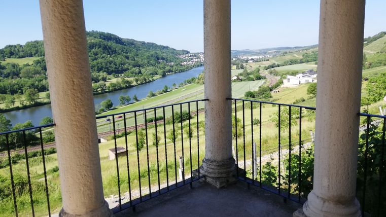 Ein Balkon mit Säulen bietet einen Blick auf einen Fluss und grüne Landschaften. Der Himmel ist klar und die Umgebung ist ruhig und malerisch.