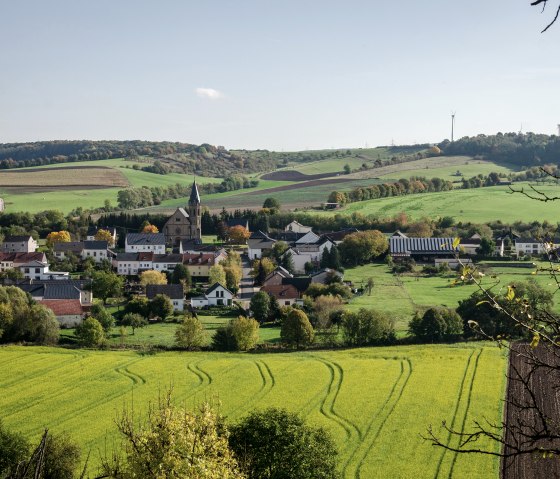 Panoramablick auf das Dorf Alsdorf mit einer Kirche im Zentrum, umgeben von gr&uuml;nen Feldern und H&uuml;geln unter blauem Himmel., &copy; Tourist-Info Bitburger Land