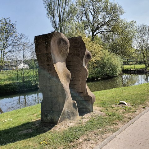 Stone sculpture on the riverbank, surrounded by green landscape and trees. A clear sky and a path are also visible., © TI Bitburger Land, Steffi Wagner