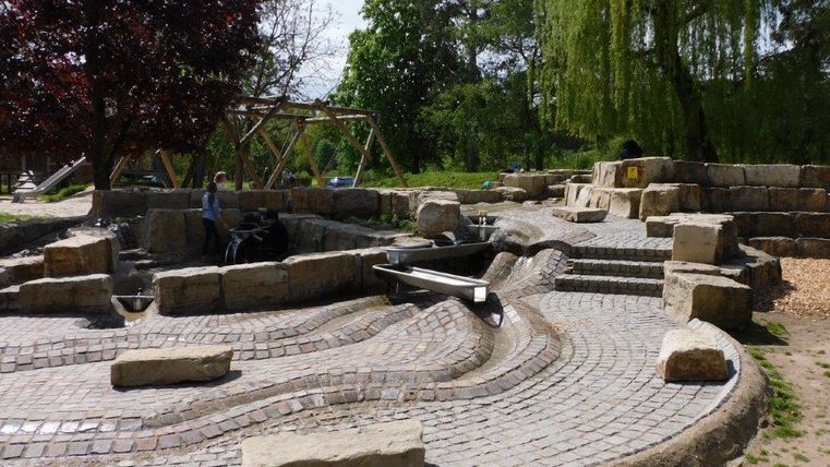 Ein Spielplatz mit natürlichen Steinen und Wasserkanälen. Kinder spielen und erkunden die unterschiedlichen Bereiche der Anlage.