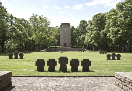 Un monument de guerre avec des croix en pierre sur une prairie, une tour en arrière-plan, entourée d'arbres verts sous un ciel bleu., © TI Bitburger Land