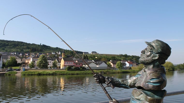 Eine Bronze-Skulptur eines anglernden Mannes steht an einem Fluss. Im Hintergrund sind malerische Häuser und sanfte Hügel zu sehen.