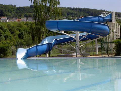 Eine blaue Rutsche neben einem klaren Schwimmbecken. Im Hintergrund sind Bäume und Hügel zu sehen.