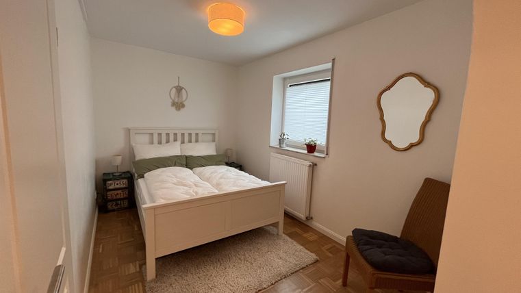 Ein gemütliches Schlafzimmer mit einem weißen Bett und einer Holzlampe. Neben dem Bett steht ein Stuhl und es gibt ein Fenster mit einem kleinen Tisch.