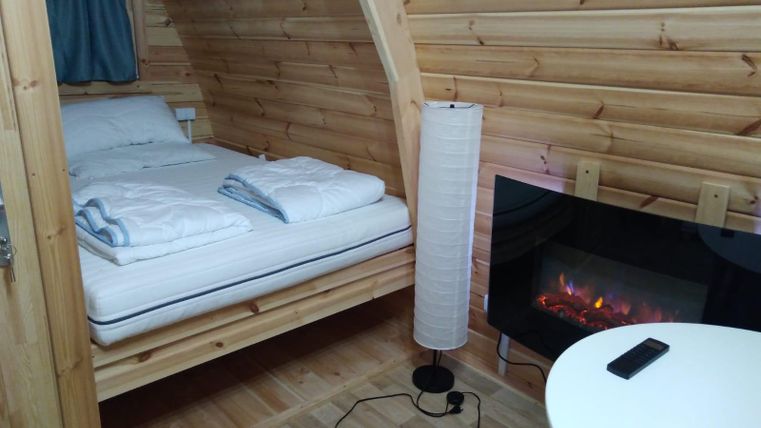 Ein gemütliches Zimmer aus Holz mit einem Bett, einer Stehlampe und einer elektrischen Heizung. Die Wände sind in warmen Holztönen gehalten, und es gibt einen kleinen Tisch.