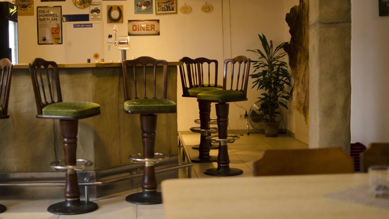 Eine gemütliche Bar mit mehreren Hockern und einer rustikalen Inneneinrichtung. Im Hintergrund sind dekorative Schilder und Pflanzen zu sehen.