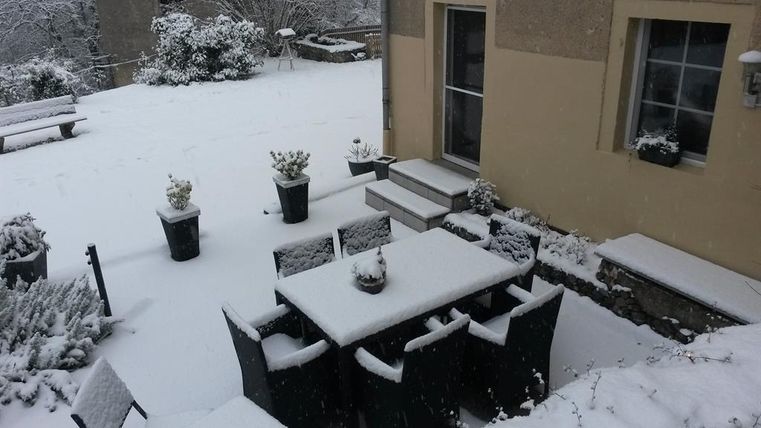 Ein verschneiter Garten mit einem Tisch und Stühlen, die mit Schnee bedeckt sind. Im Hintergrund sind ein paar Pflanzen und ein Haus sichtbar.