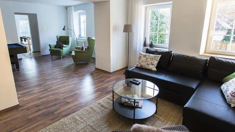 Ein modernes Wohnzimmer mit einer gemütlichen Couch und einem Tisch. Im Hintergrund sind grüne Sessel und ein heller Raum zu sehen.