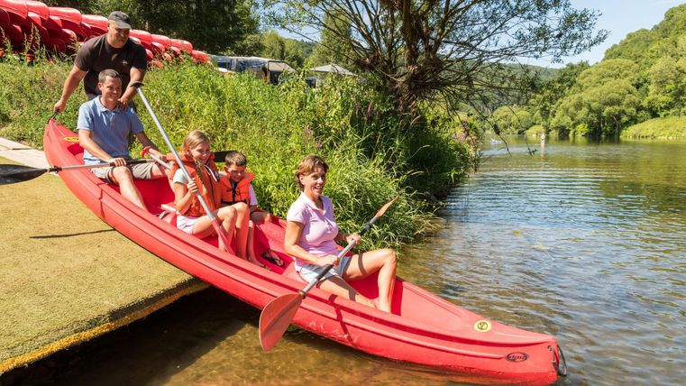 Eine Familie paddelt in einem roten Kajak auf einem ruhigen Gewässer. Im Hintergrund sind Bäume und ein sonniger Himmel zu sehen.