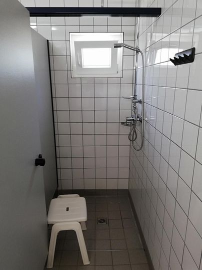 Eine einfache Duschkabine mit weißen Fliesen und einem Fenster. Ein Plastiksitz steht im Raum.