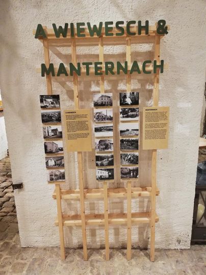 Eine Ausstellungstafel mit dem Titel „A Wiewesch & Manternach“. Sie zeigt historische Fotos und Informationen auf gelben Plaketten.