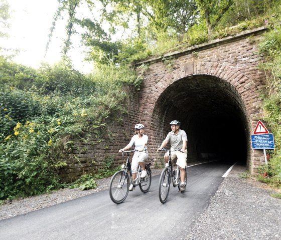 Two cyclists ride out of an overgrown tunnel on the Vennbahn near Lommersweiler. There is a warning sign at the edge., © vennbahn.eu