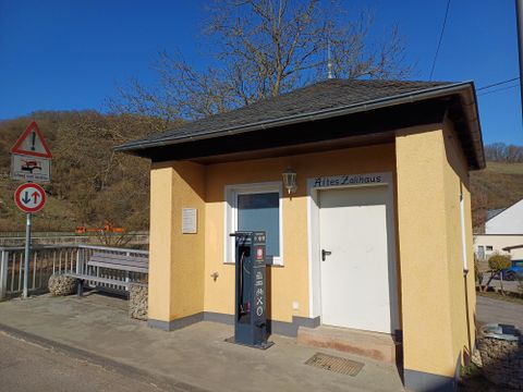 Ein kleines, gelbes Gebäude mit einem schwarzen Dach steht an einer Straße. Es trägt den Schriftzug "Altes Zollhaus" und hat einen Parkplatz mit einer alten Zapfsäule.