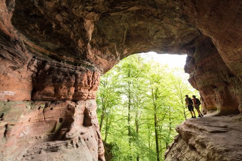Eine beeindruckende Höhle mit roten Felsen und einem Blick auf einen grünen Wald. Zwei Wanderer stehen am Höhlenausgang und genießen die Aussicht.