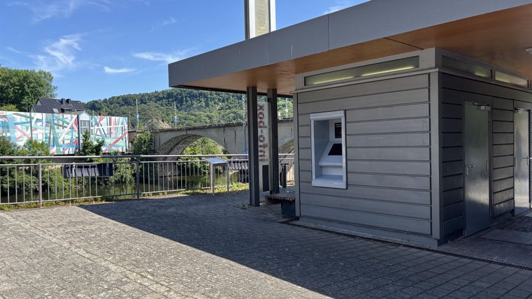 Ein moderner Kiosk aus Holz und Glas steht auf einem gepflasterten Platz. Im Hintergrund ist eine Brücke und farbenfrohe Graffiti zu sehen.