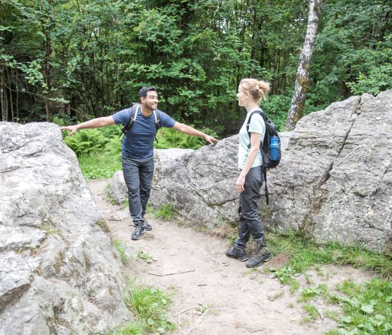Deux randonneurs se tiennent entre de gros rochers dans la forêt et discutent. Ils portent des sacs à dos et des vêtements de randonnée., © Eifel-Tourismus GmbH, shapefruit AG A. Röser