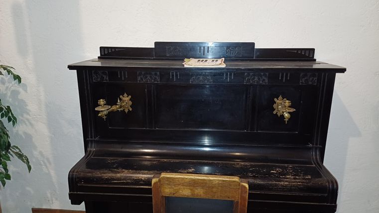 Ein schwarzes Klavier mit dekorativen Details steht an der Wand. Vor dem Klavier befindet sich ein einfacher Holzstuhl.