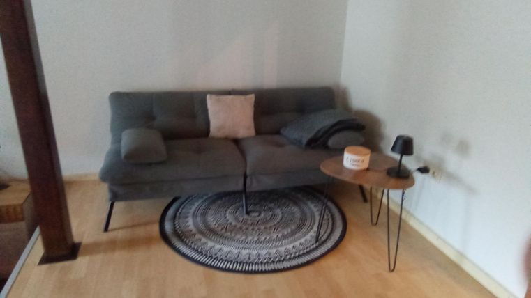 Ein modernes Wohnzimmer mit einem grauen Sofa und einem runden Teppich. Neben dem Sofa steht ein kleiner Tisch mit einer Lampe und einem Deko-Element.